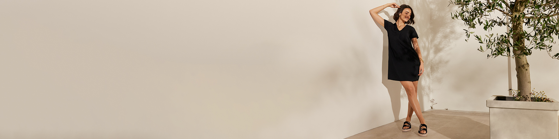En kvinde i sort kjole og sandaler danser afslappet foran en lys væg og et oliventræ.