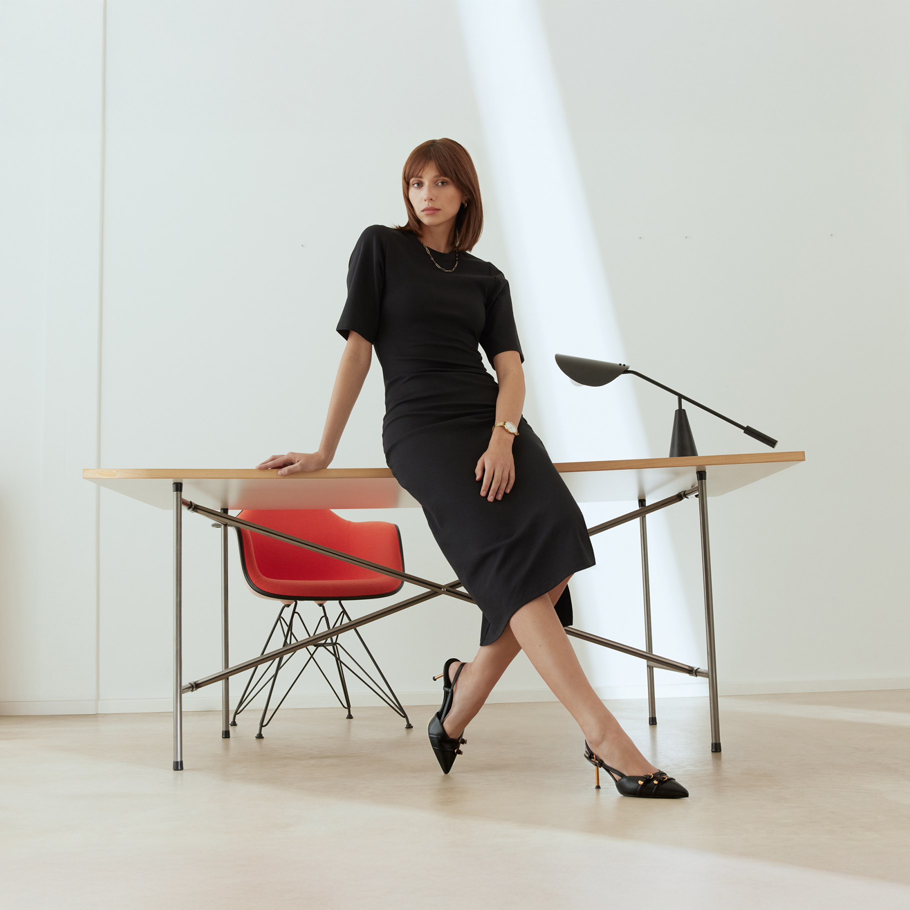 Elegantn&iacute; žena v čern&yacute;ch &scaron;atech se op&iacute;r&aacute; o designov&yacute; stůl s červenou židl&iacute; v minimalistick&eacute;, světl&eacute; kancel&aacute;ři.