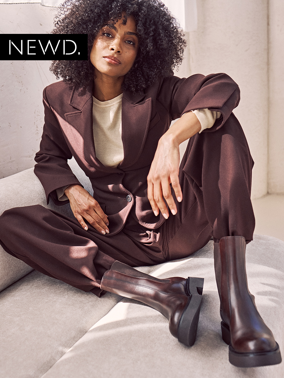 Modisches Outfit mit braunem Anzug, cremefarbenem Pullover und kniehohen Lederstiefeln, auf hellem Sofa fotografiert. NEWD-Logo sichtbar.