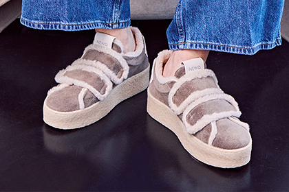 Füße in Jeans und beigen Sneakers mit Fell, die auf dunklem Untergrund stehen.