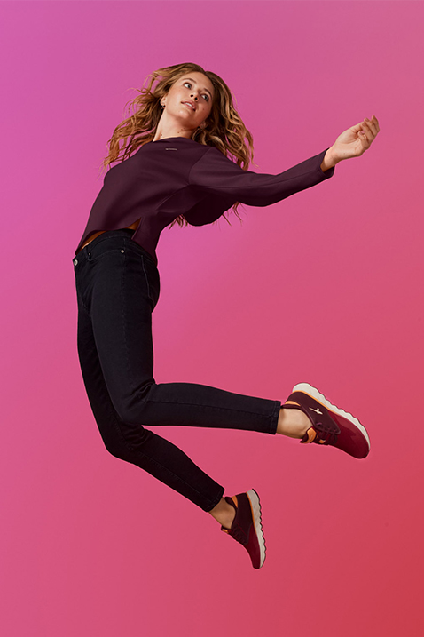 Eine dynamische Tänzerin in schwarzer Kleidung springt elegant vor einem leuchtend rosa Hintergrund mit dunkelroten Light Sneakern.