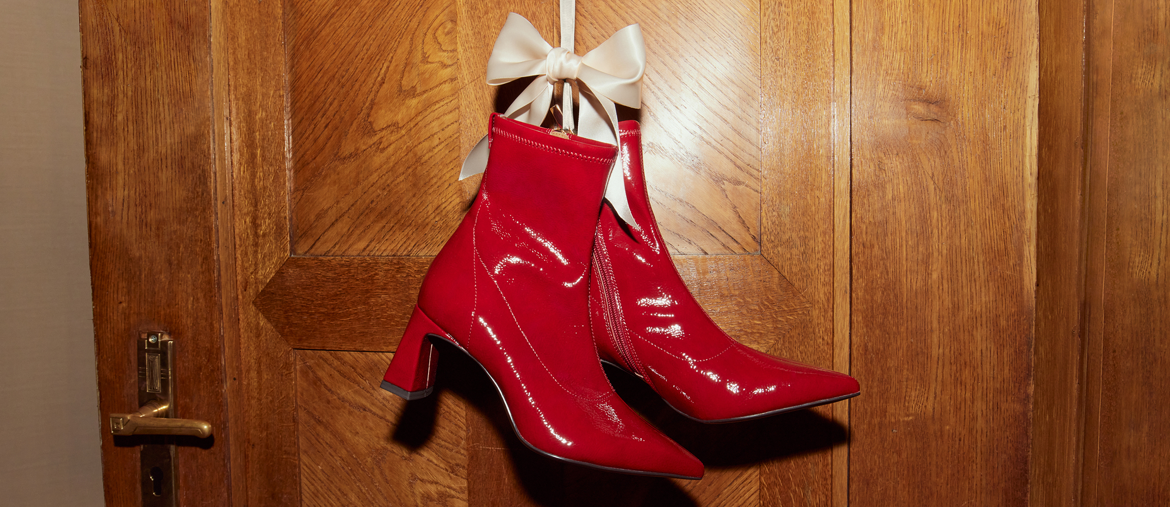 Shiny red patent leather boots with white bows hang on a wooden wall as a festive Christmas decoration.