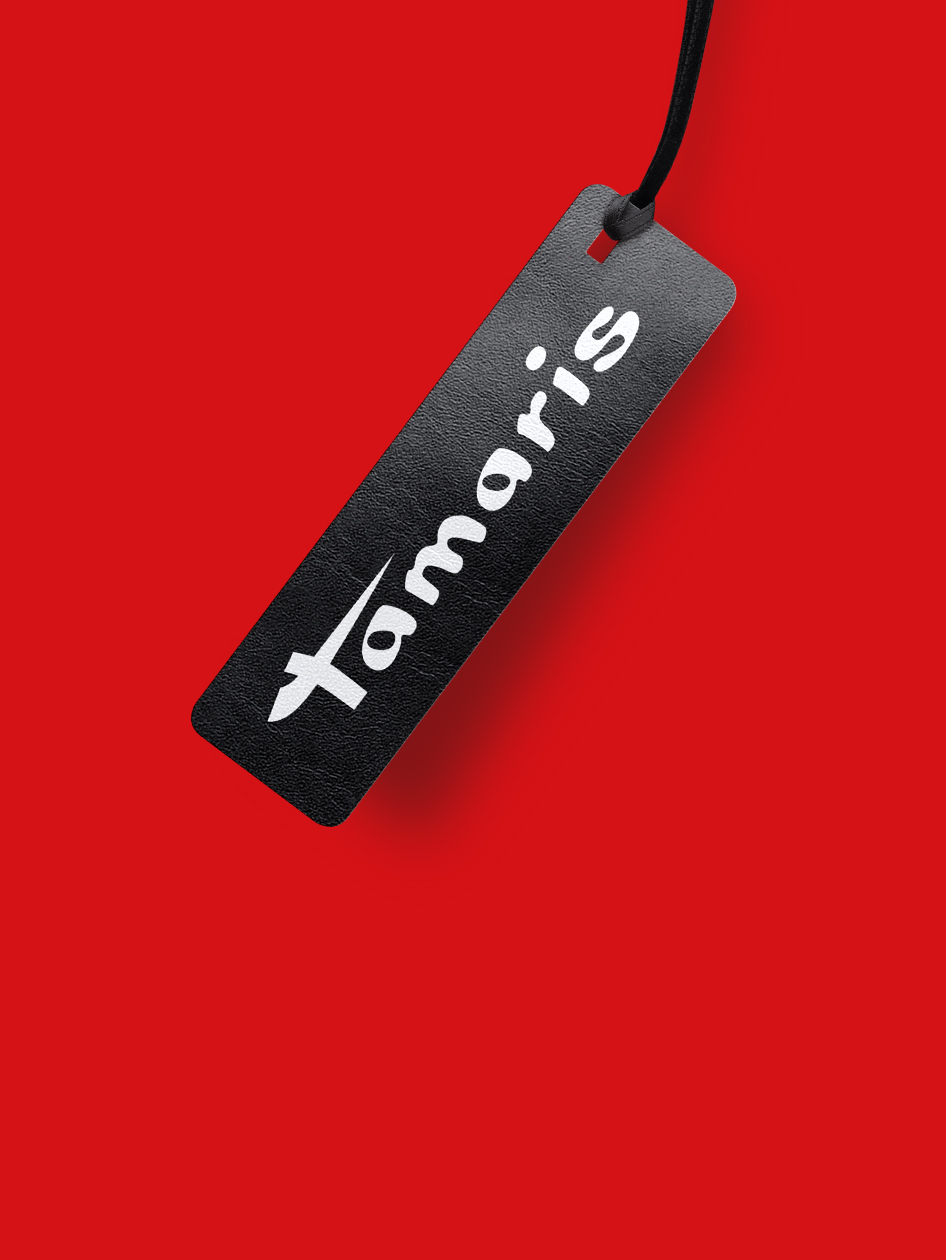 Schwarzes Tamaris-Hangtag mit wei&szlig;em Logo, an schwarzer Schnur h&auml;ngend, vor rotem Hintergrund.