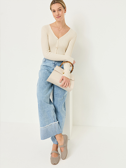 Woman in a cream cardigan, wide light blue jeans and grey strap ballerinas, holding a cream handbag.