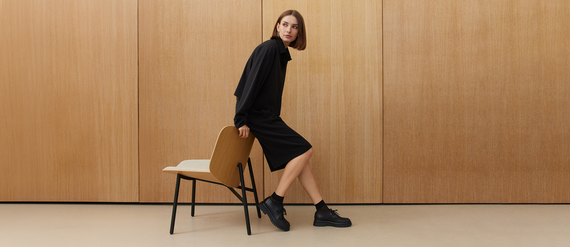Woman in a black outfit and black lace‑up shoes leaning against a wooden chair in front of a light wooden wall.