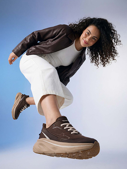 Woman with curly hair in a brown leather jacket, white trousers and brown sneakers strikes a dynamic pose against a blue background.