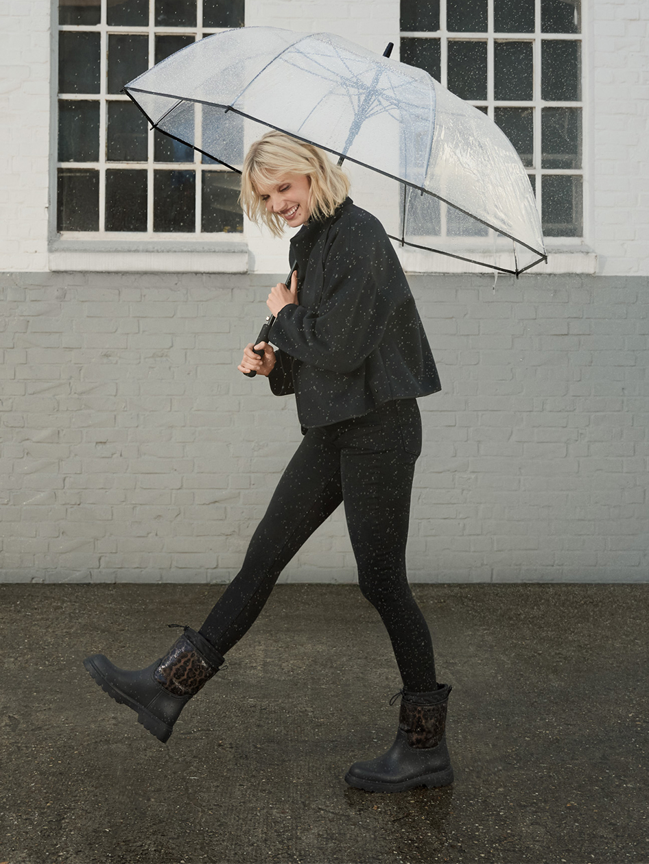 Une personne en tenue noire avec un parapluie transparent danse joyeusement sous la pluie avec des bottes en caoutchouc devant un mur de briques blanches.