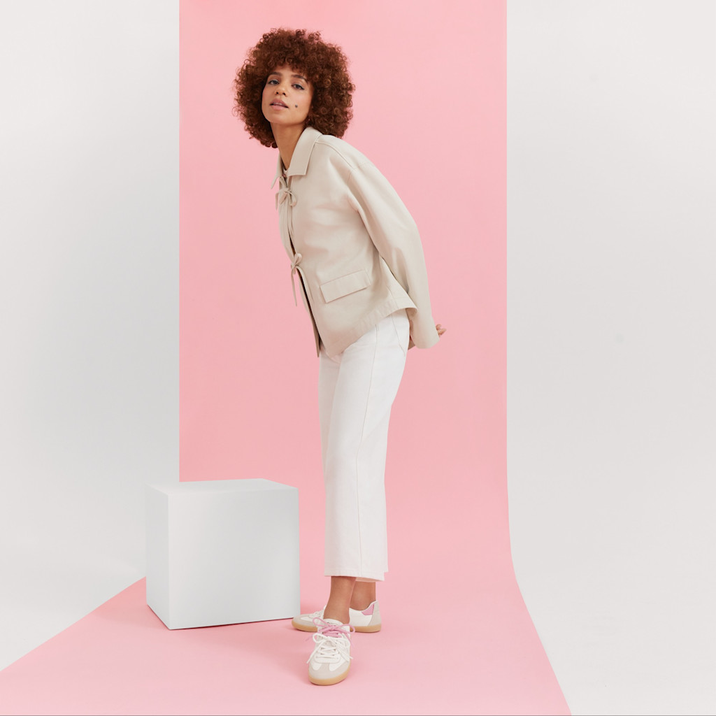 Modello in blazer beige e pantaloni bianchi davanti a uno sfondo rosa con sneakers bianche in posa casual.