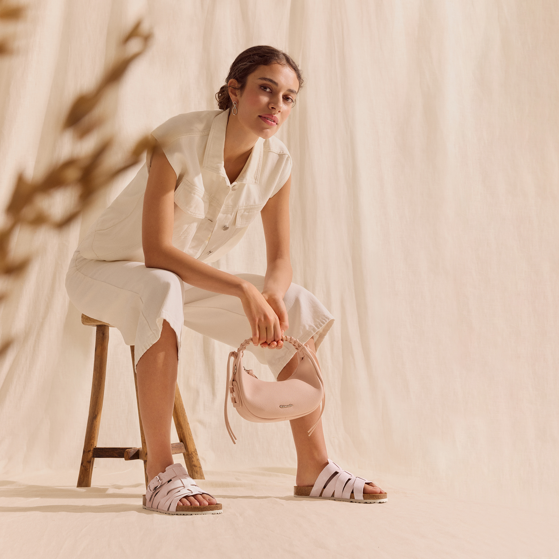 Vrouw in een beige, zomerse outfit zit op een kruk, draagt roze sandalen en houdt een kleine roze handtas vast.