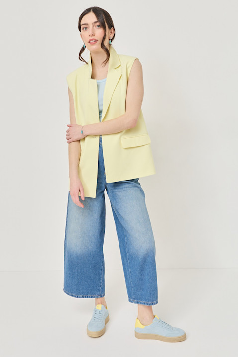Model with curly hair in a cream-colored outfit and sneakers poses in front of a pink background next to a white pedestal.