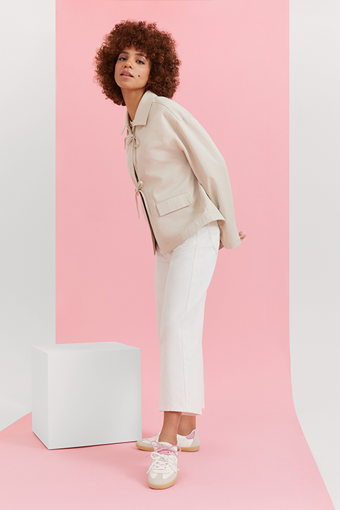 Mannequin aux cheveux boucl&eacute;s, en tenue cr&egrave;me et baskets, posant devant un fond rose &agrave; c&ocirc;t&eacute; d&rsquo;un socle blanc.