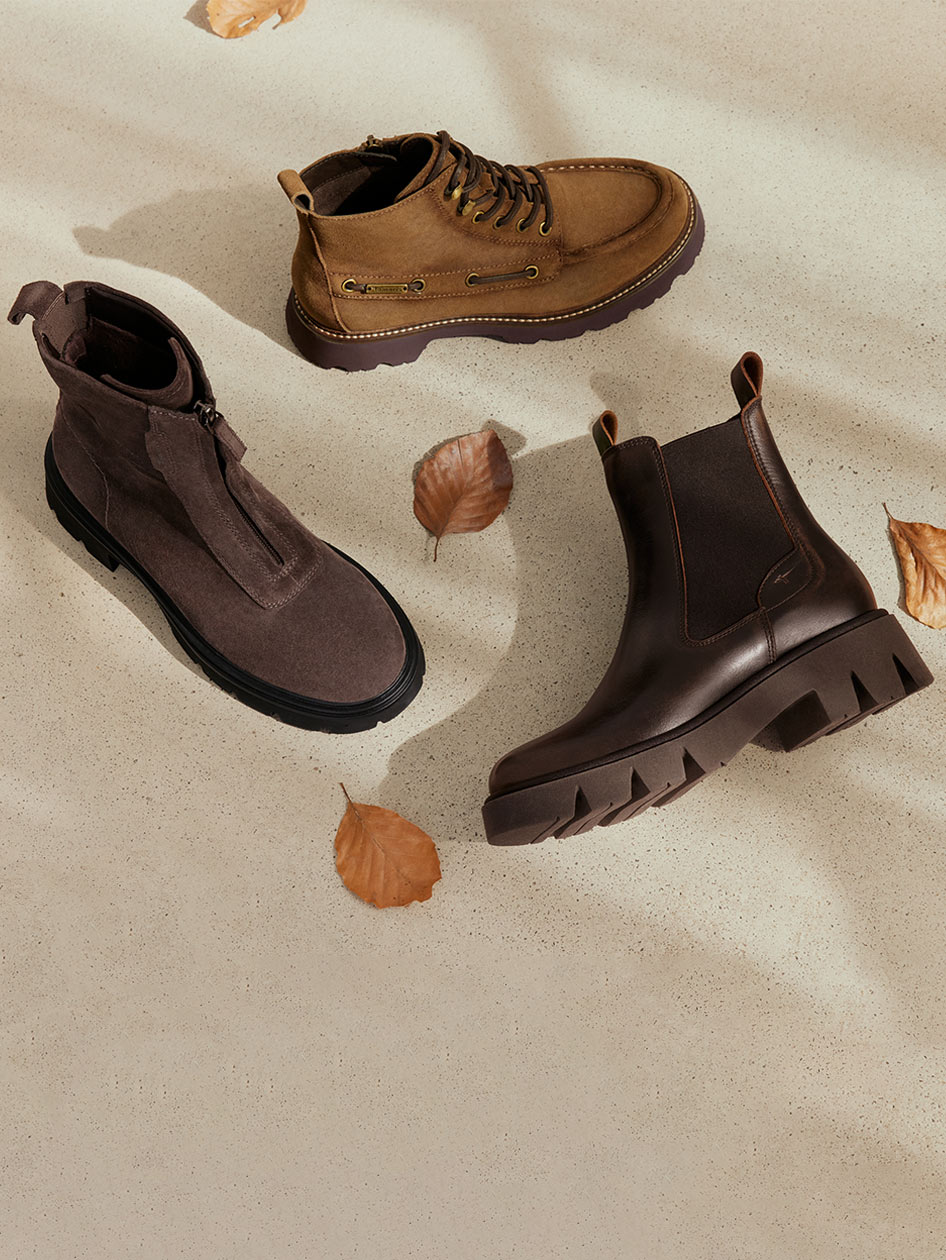 Drei Herbststiefel in Braun- und Graut&ouml;nen auf hellem Untergrund, umgeben von trockenen Herbstbl&auml;ttern.