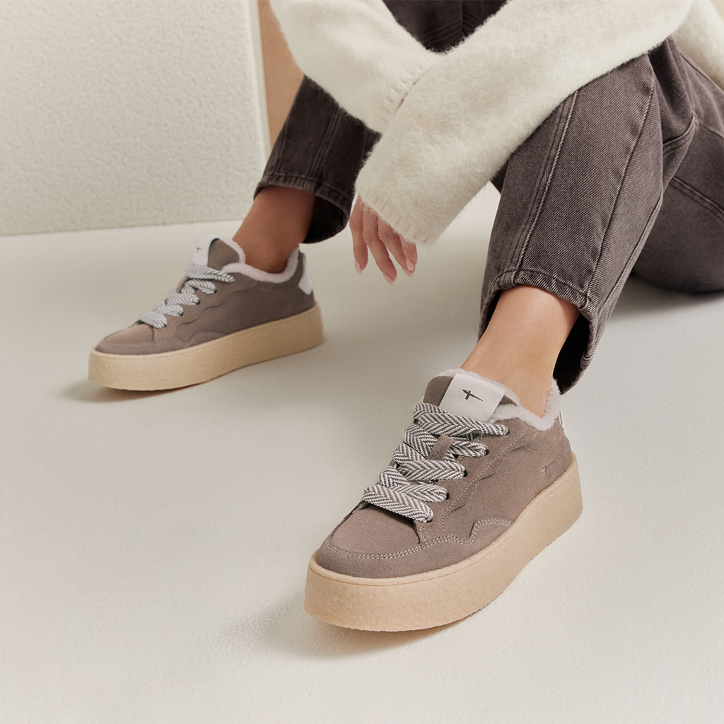 Femme portant des sneakers &agrave; plateau taupe avec bord en fourrure et lacets &agrave; motifs, associ&eacute;es &agrave; un jean gris et un pull cr&egrave;me.