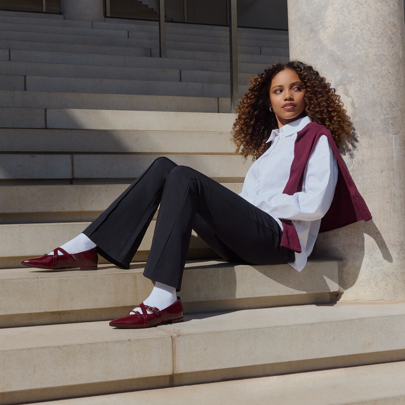 A person in elegant business clothes is sitting relaxed on a wide stone staircase, wearing a wine-red vest and matching loafers.