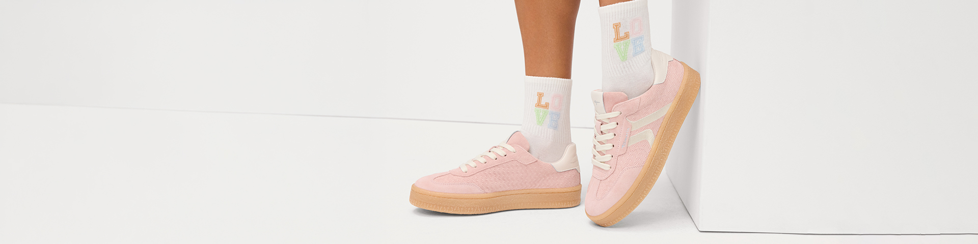Pink sneakers with rubber soles and white socks with a colorful LOVE print on women&rsquo;s legs against a white background.