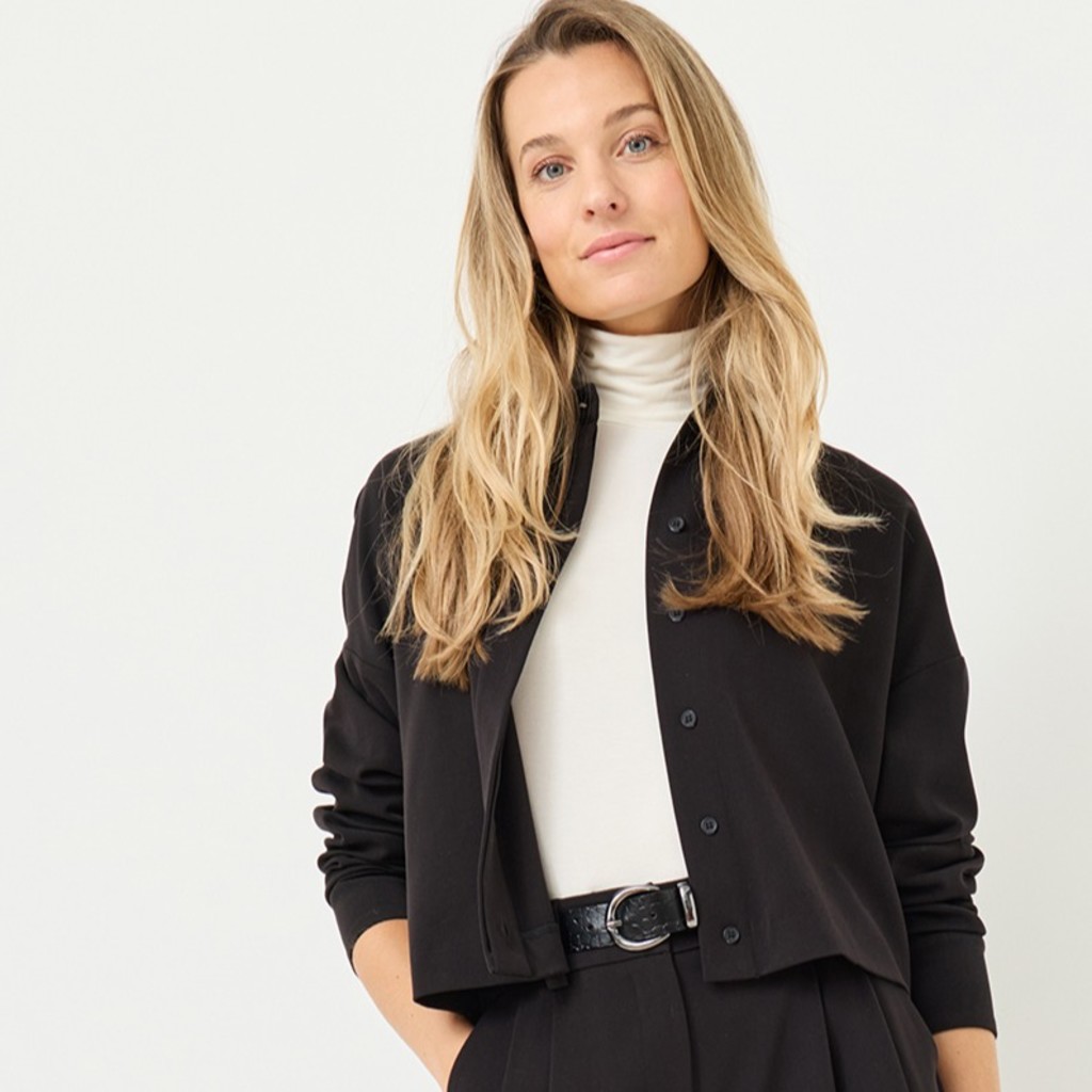 Woman in elegant business attire with black jacket and white turtleneck sweater against a light background.