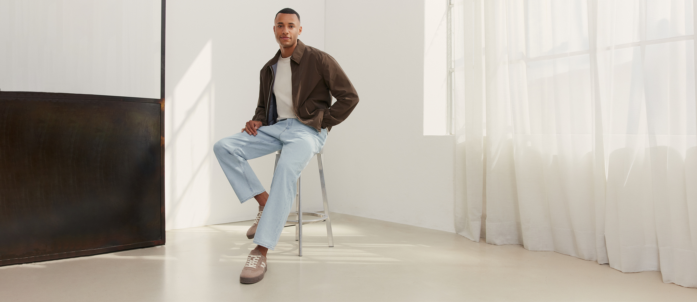 Man sitting on a stool, wearing a brown leather jacket, light-coloured t-shirt, light blue jeans and beige trainers in a bright, modern room.