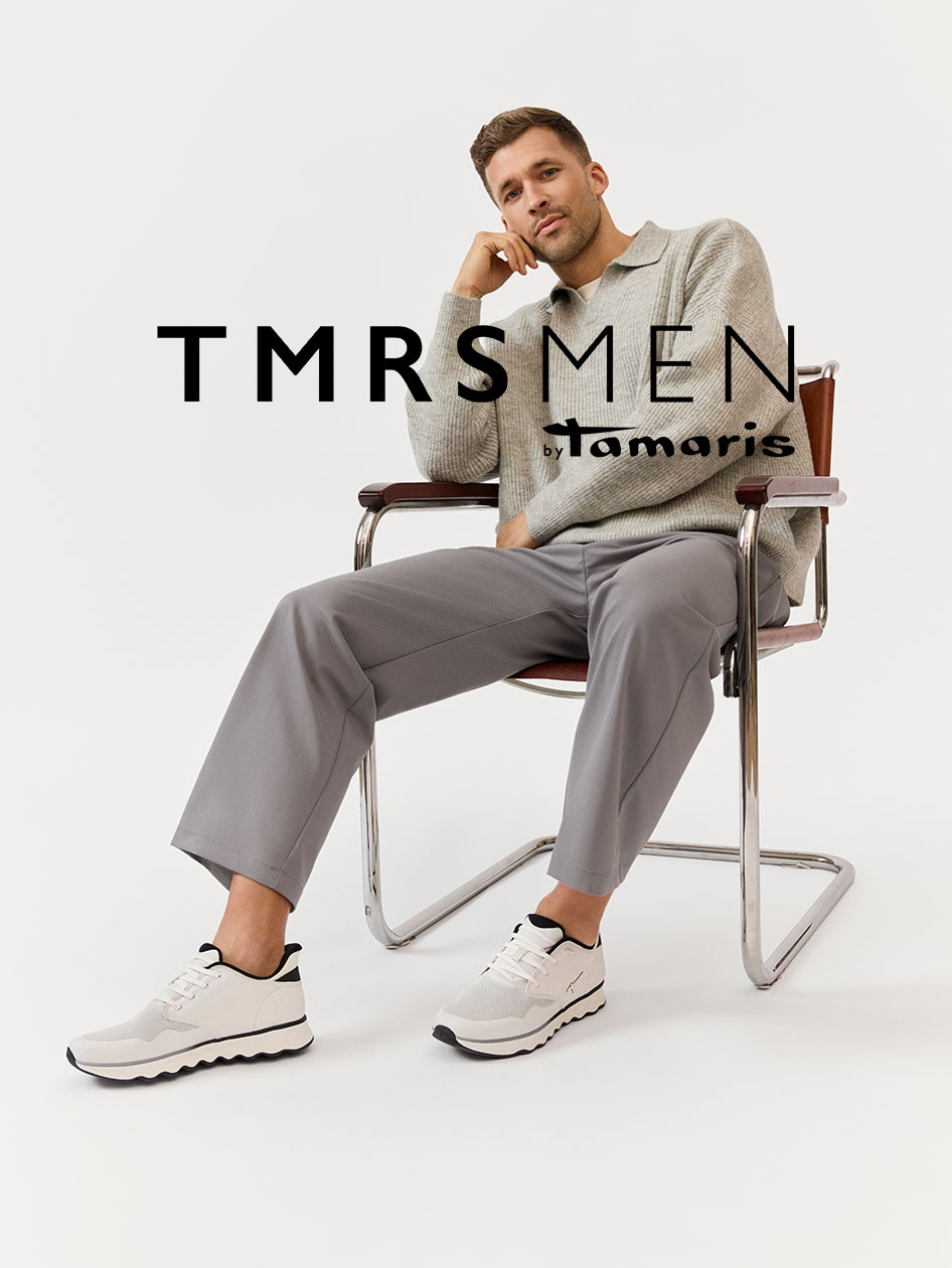 Model v b&eacute;žov&eacute;m svetru a &scaron;ed&yacute;ch kalhot&aacute;ch sed&iacute; uvolněně v designov&eacute; židli a m&aacute; na sobě b&iacute;l&eacute; tenisky TMRS MEN od Tamaris.