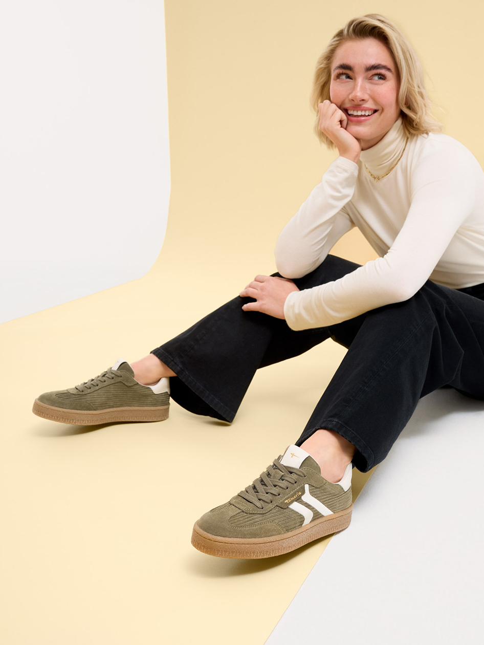 Lächelnde Person in weißem Rollkragenpullover und schwarzer Hose, trägt olivgrüne Sneaker mit weißen Streifen, auf beigem Hintergrund.