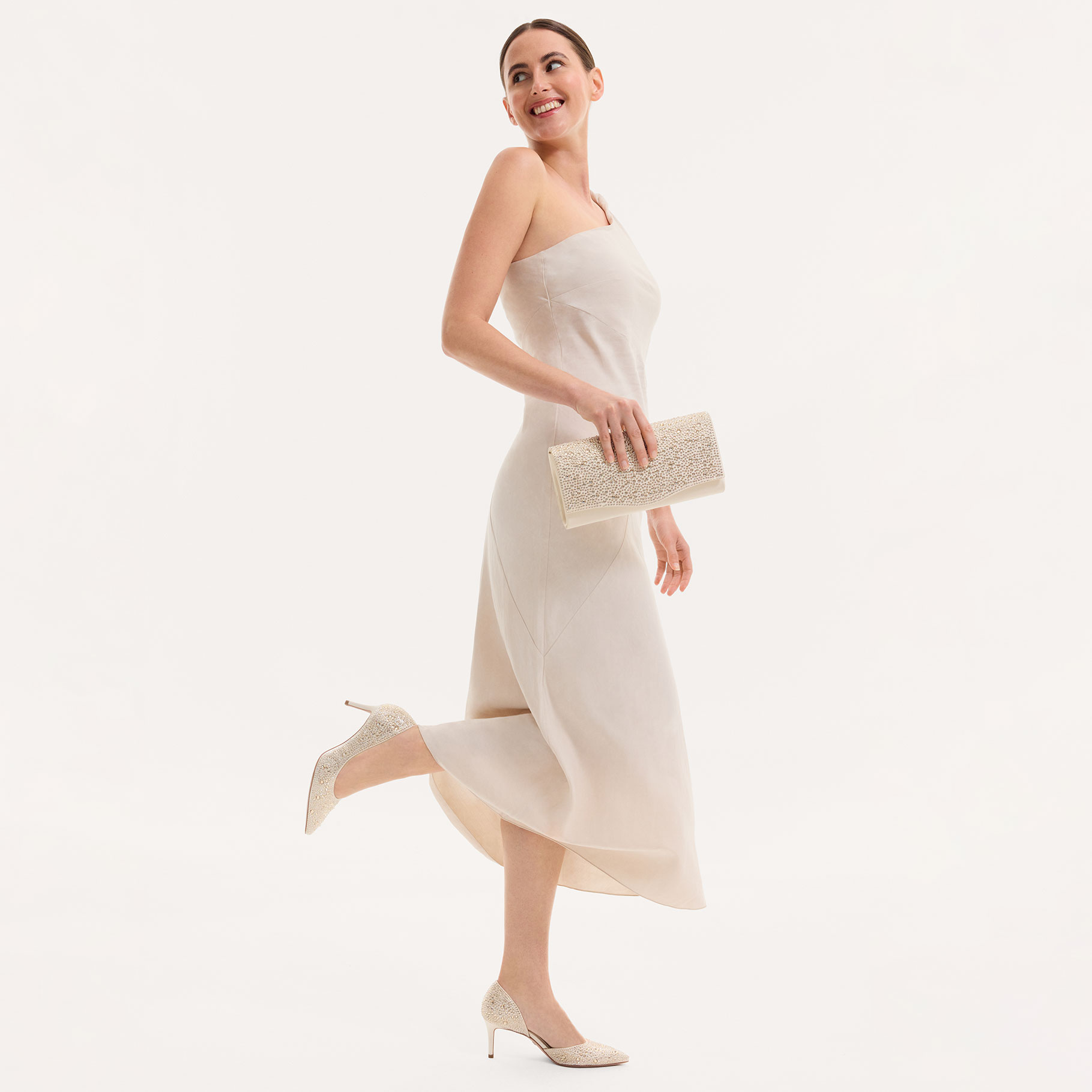 Smiling woman in an elegant white evening dress with a sparkling clutch and matching stilettos.