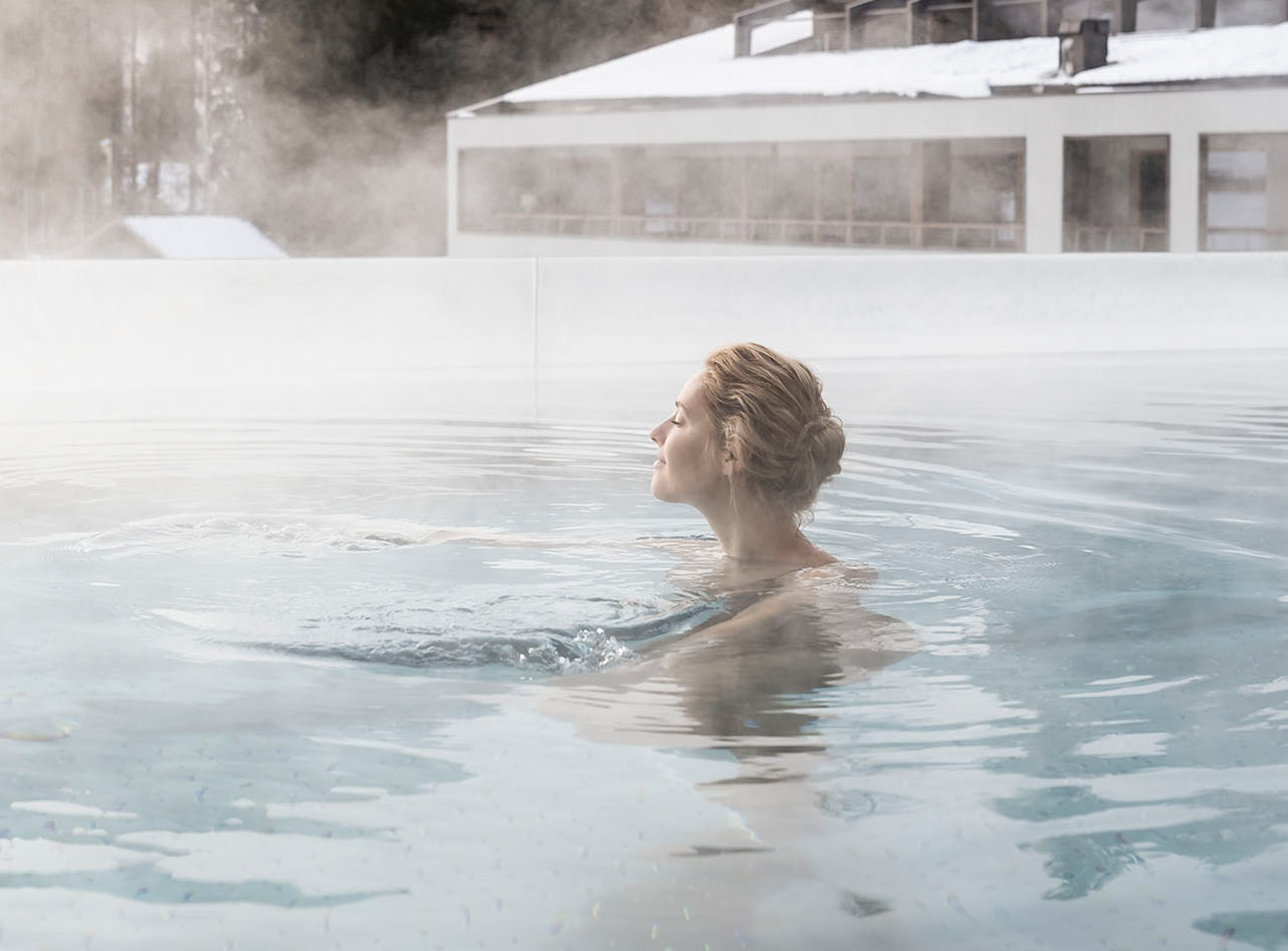 Frau entspannt im dampfenden Au&szlig;enpool eines Spa-Hotels im Winter, mit schneebedecktem Dach im Hintergrund.