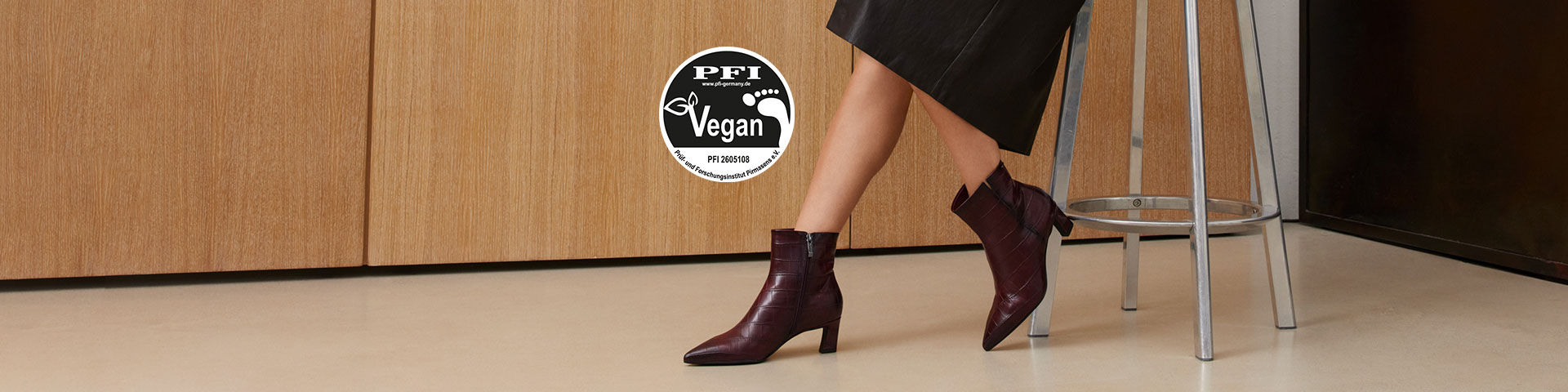 Kobiece nogi w ciemnoczerwonych wegańskich botkach na obcasie, czarne spodnie, certyfikat PFI Vegan, drewniana ściana w tle.