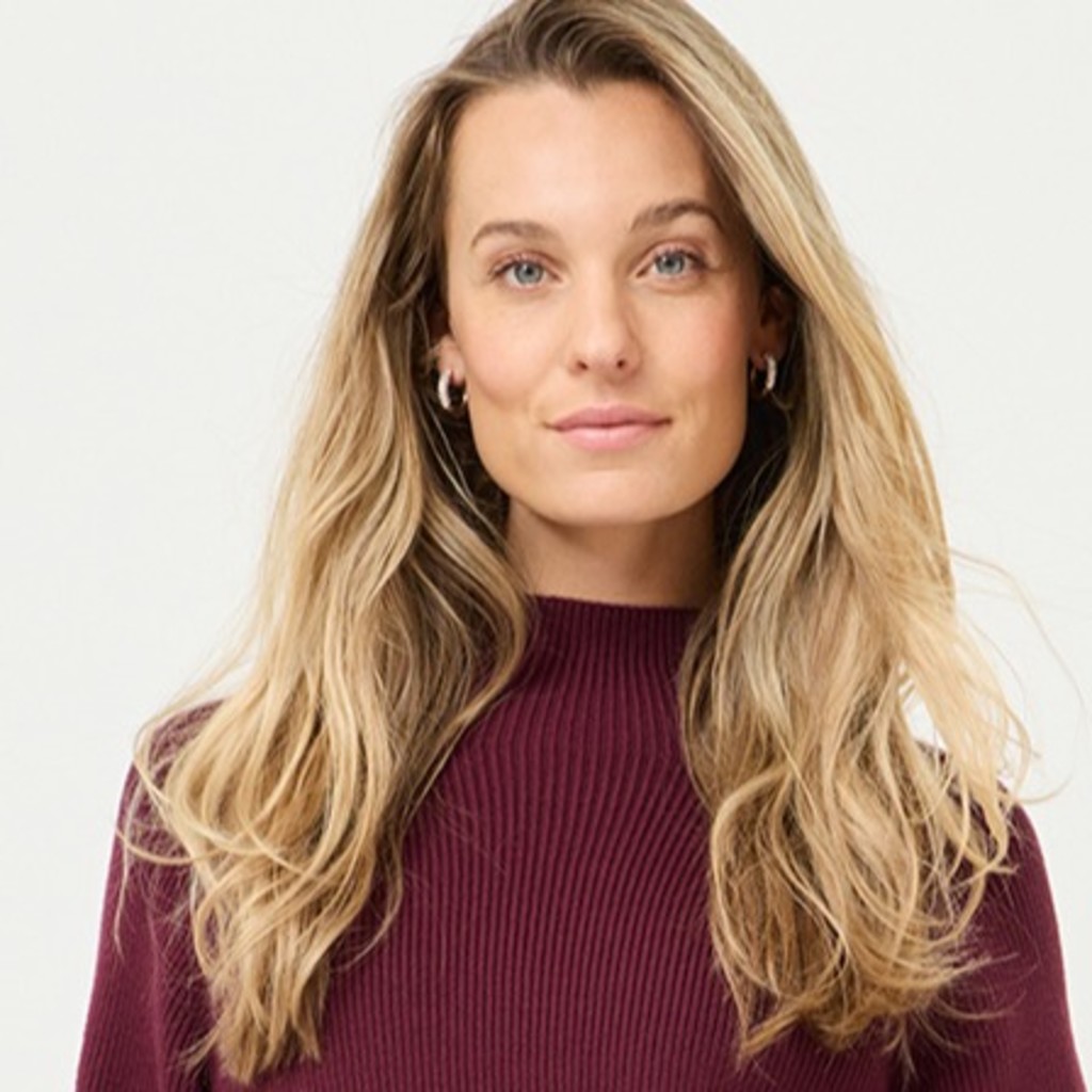 Eine junge Frau mit langen blonden Locken tr&auml;gt einen weinroten Pullover vor hellem Hintergrund.