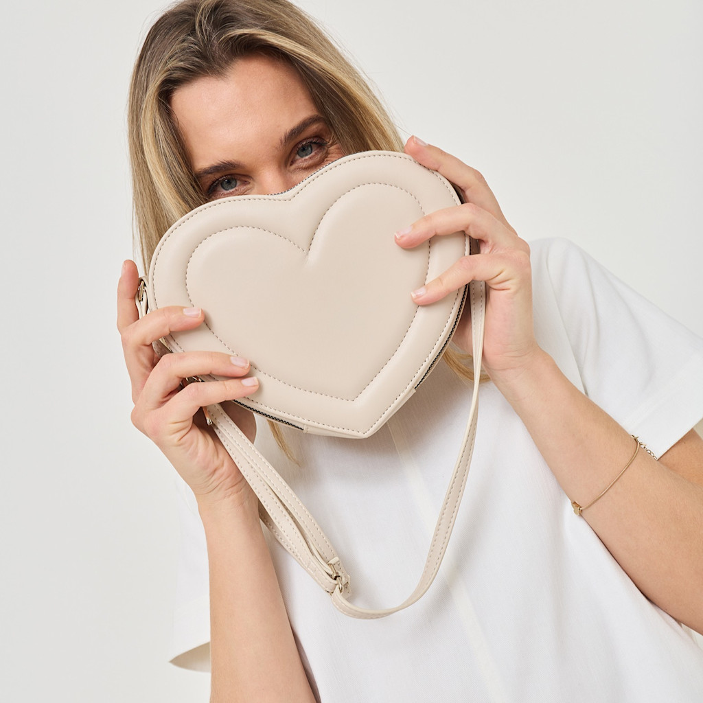 A beige heart-shaped handbag is held in front of the face, with only the eyes visible above the bag.