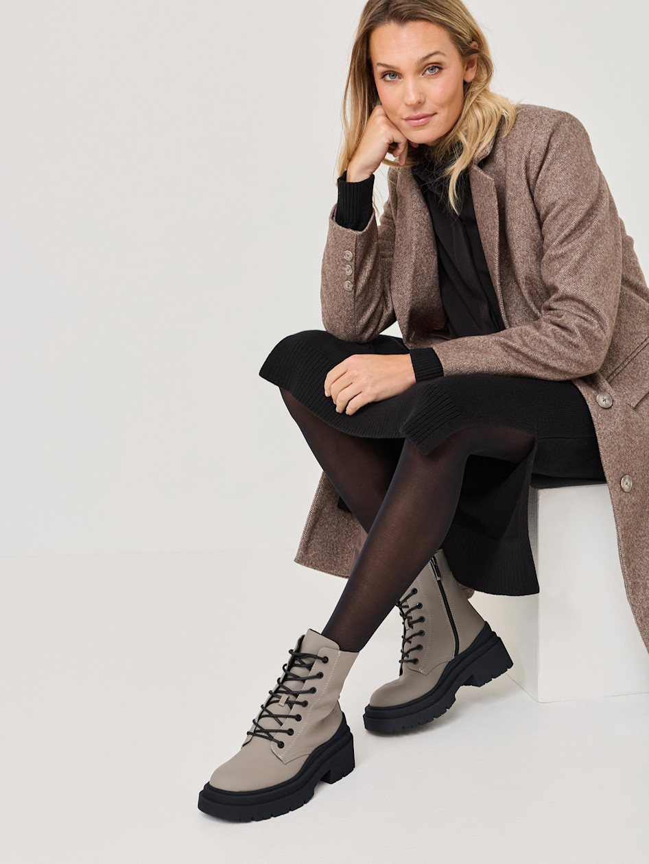 Modern h&ouml;stoutfit med brun kappa, svart kl&auml;nning och gr&aring; sn&ouml;rst&ouml;vlar, fotograferad sittande mot en vit bakgrund