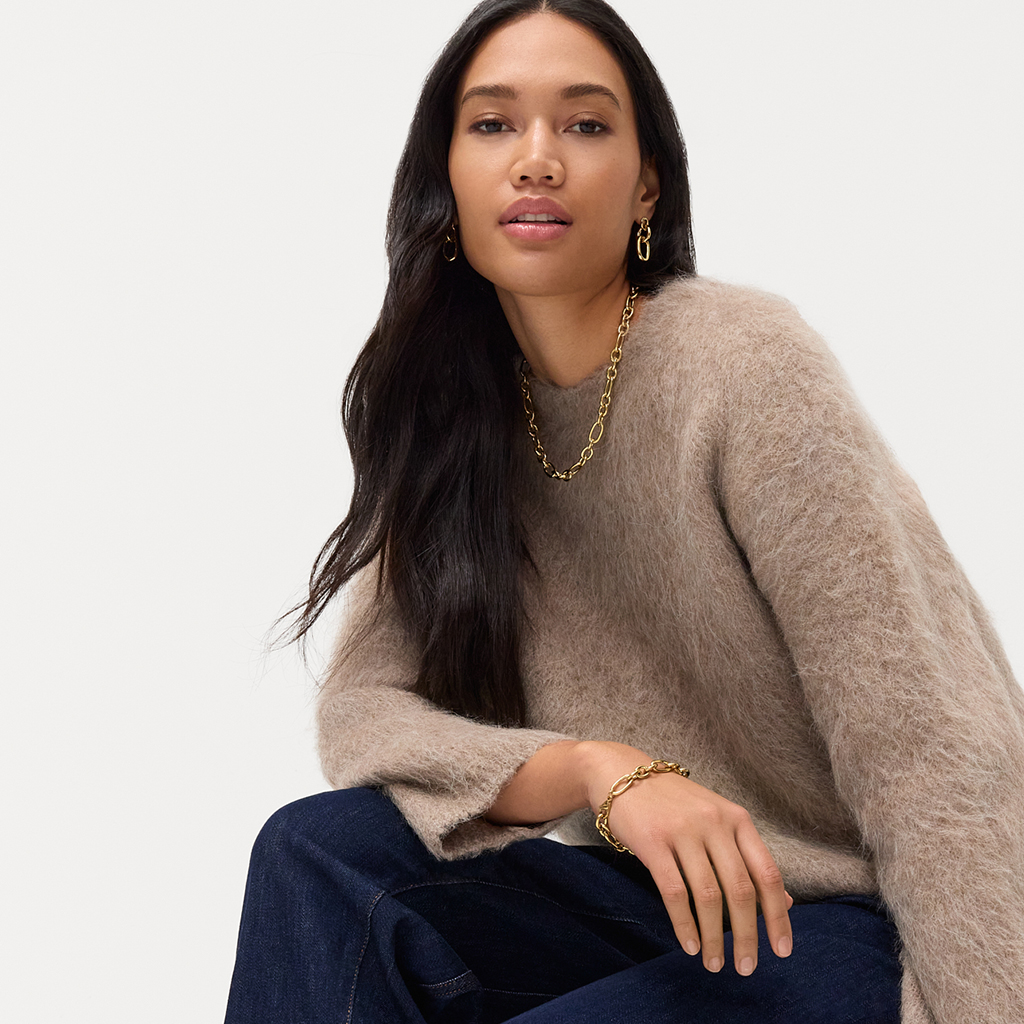 Vrouw met lang zwart haar draagt een beige fluffy trui, donkere jeans en gouden sieraden.