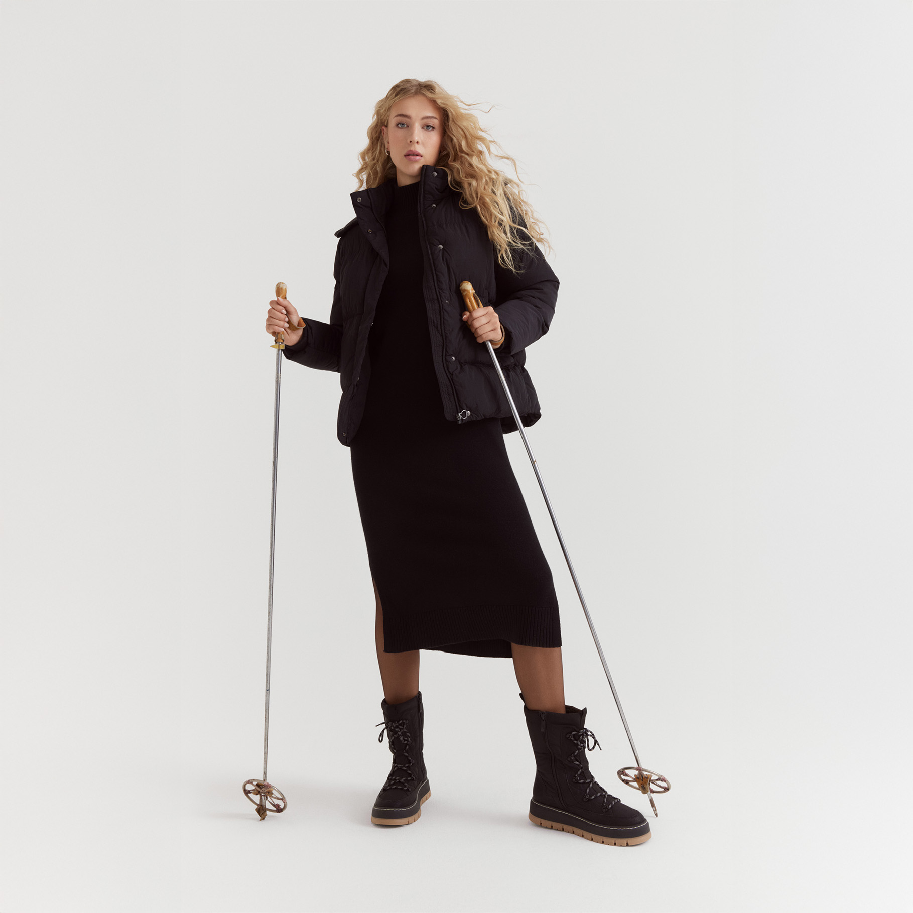 Modell in schwarzer Winterkleidung mit Skistöcken, trägt eine Daunenjacke, langes Strickkleid und robuste Schnürstiefel