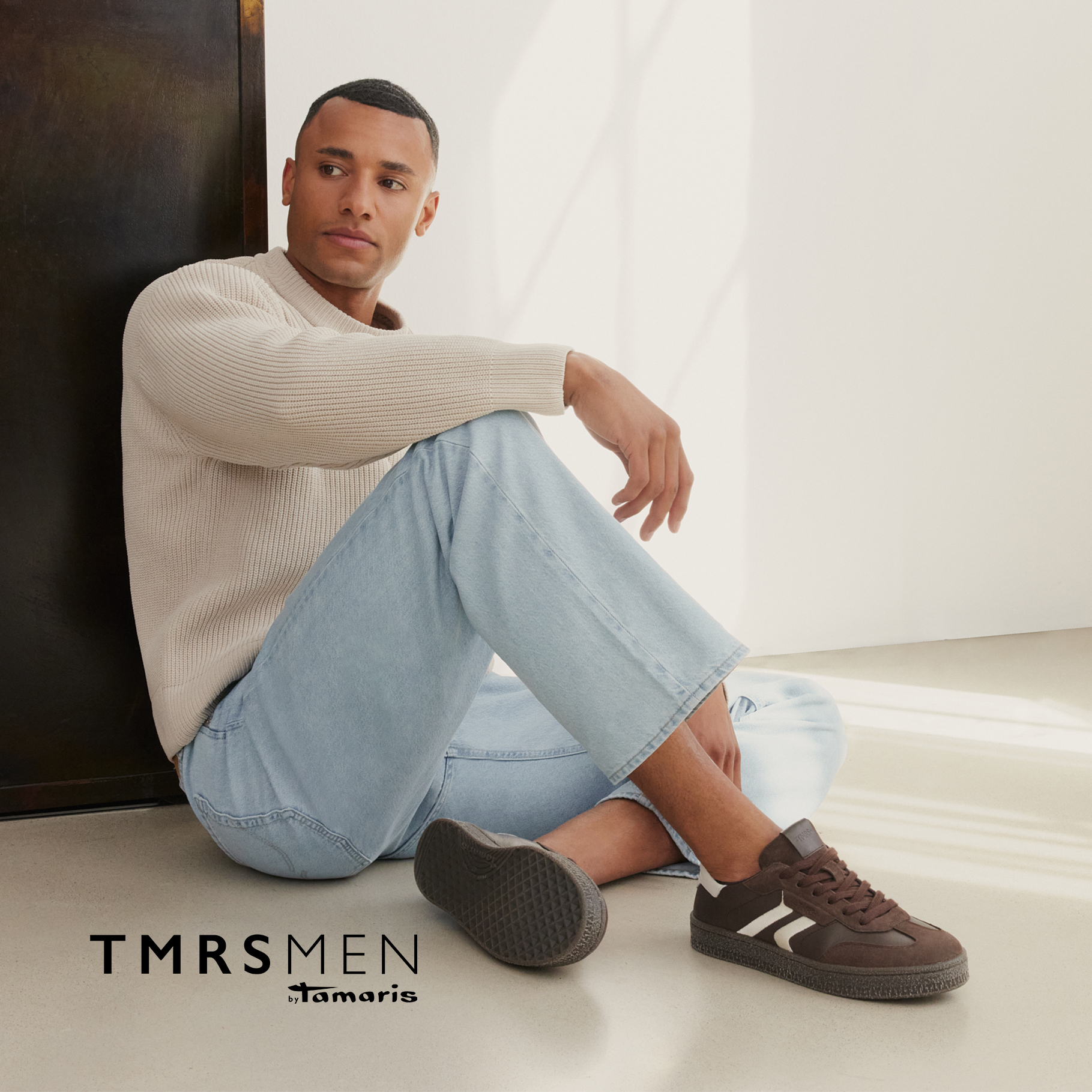 Man sitting on the floor wearing a beige knit sweater, light blue jeans and brown sneakers. Logo: TMRS MEN by Tamaris.