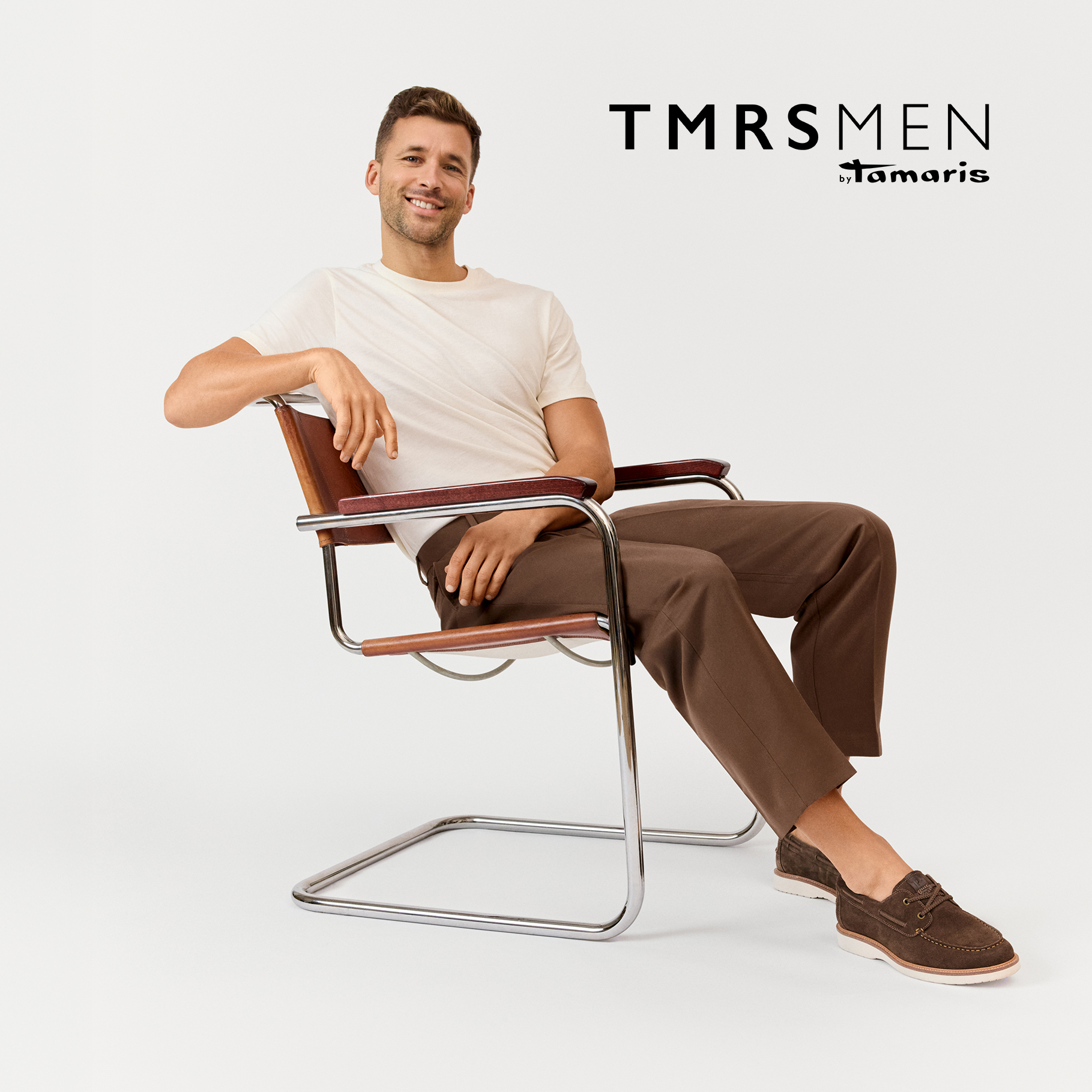 Muž v b&iacute;l&eacute;m tričku a hněd&yacute;ch kalhot&aacute;ch sed&iacute; uvolněně v designov&eacute;m křesle a m&aacute; na sobě hněd&eacute; p&aacute;nsk&eacute; boat shoes od Tamaris.