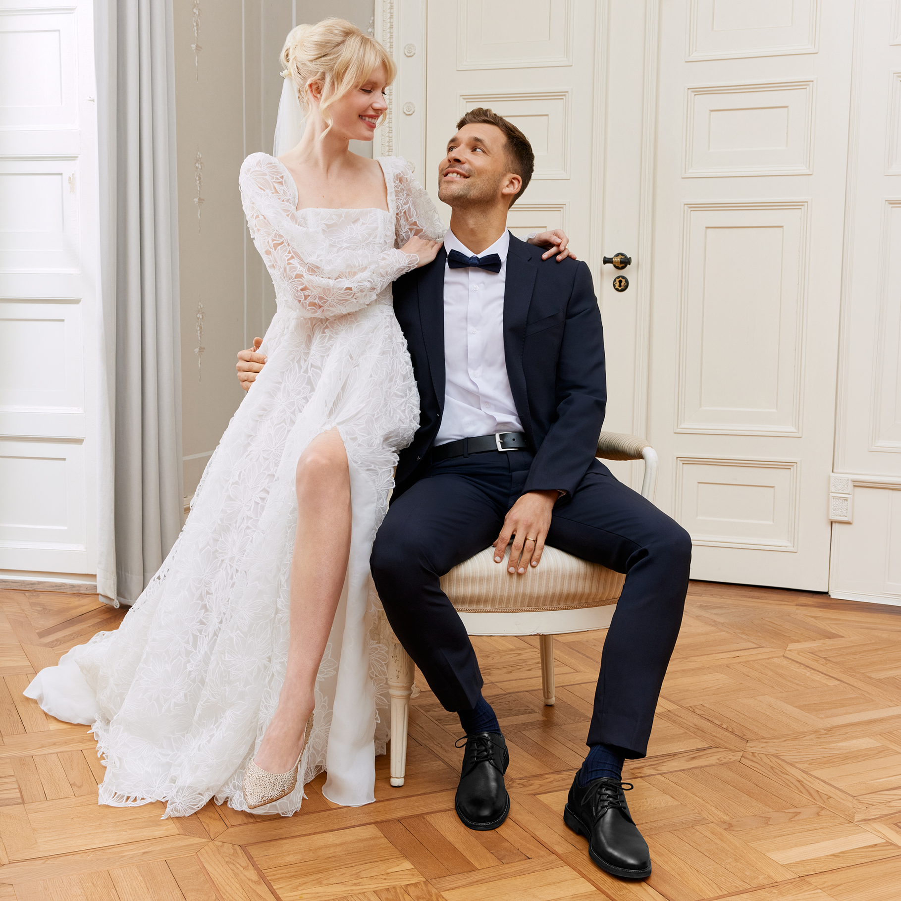 Felice coppia di sposi in abiti eleganti posa in modo giocoso su una panca antica in una stanza luminosa con pavimento in parquet.