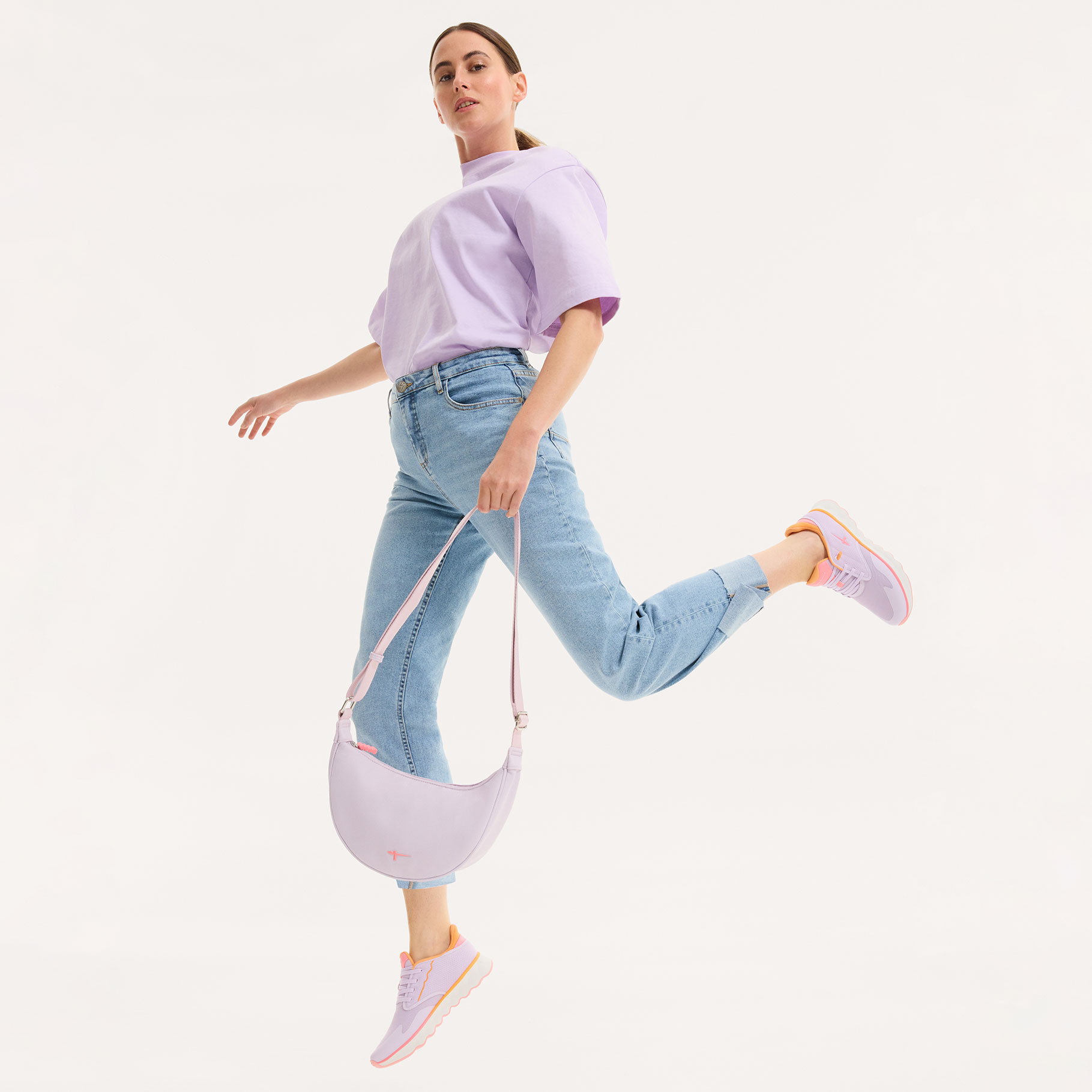 Dynamische Aufnahme einer Person in hellviolettem Oberteil und Jeans, die in pastellfarbenen Sneakers springt vor wei&szlig;em Hintergrund.