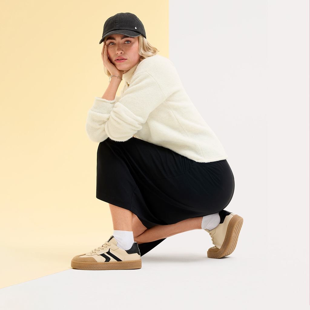 Model in sportlichem Look mit schwarzer Baseballkappe, weißem Pullover und schwarzem Rock, hockend vor beigefarbenem Hintergrund.