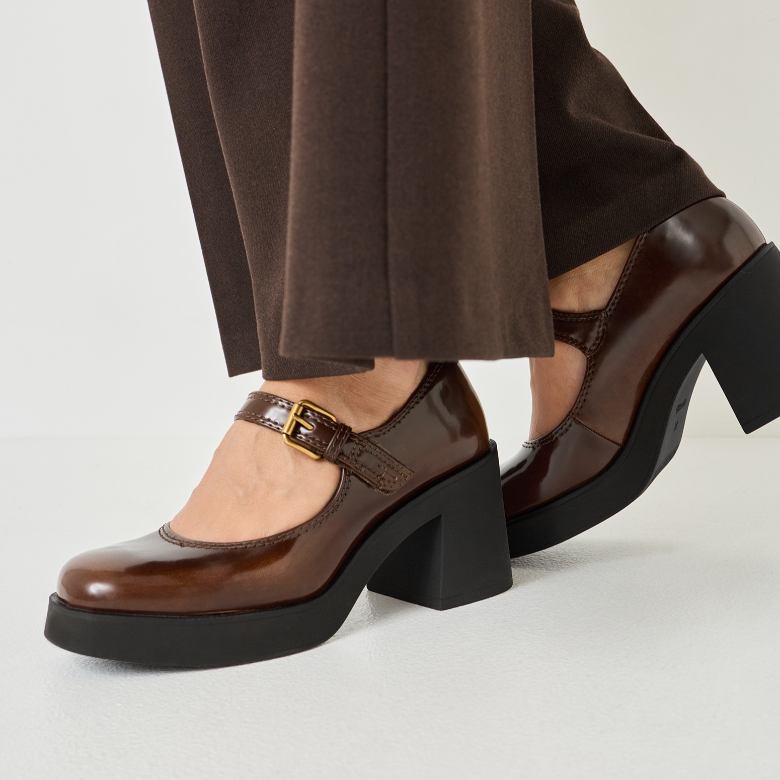 Chaussures en cuir verni marron avec lanières style Mary-Jane, semelle compensée noire et talon large, portées avec un pantalon marron.