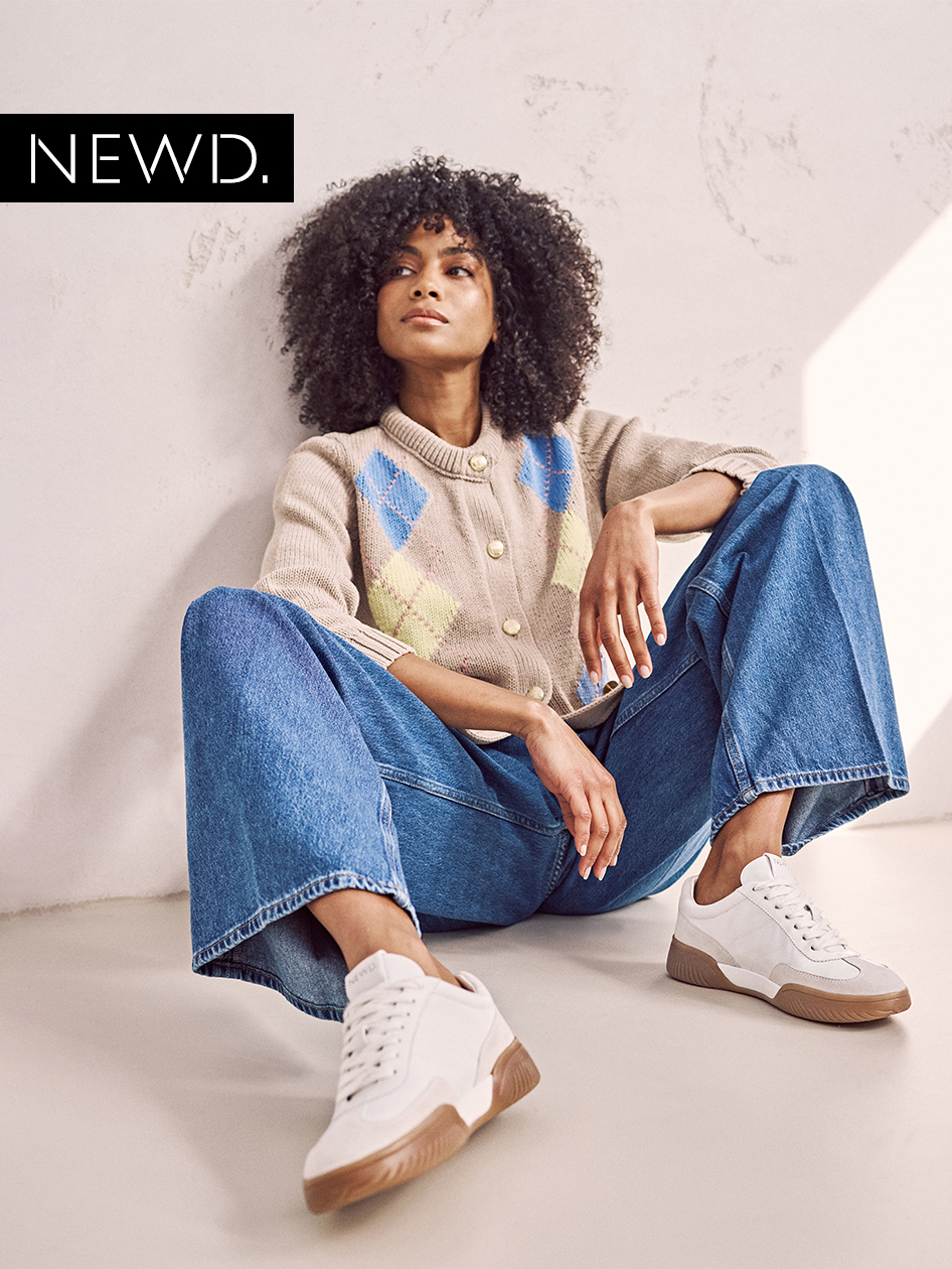 Modisches Outfit mit beigem Strickcardigan, weiter Jeans und weißen Sneakers, lässig an weißer Wand sitzend präsentiert.