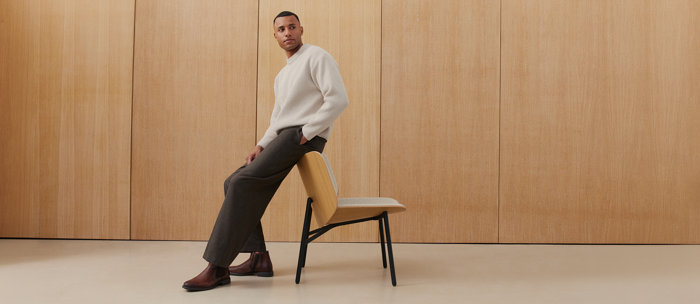 Un homme en pull cr&egrave;me, pantalon gris fonc&eacute; et bottines Chelsea marron s'appuie contre une chaise moderne devant un mur en bois.