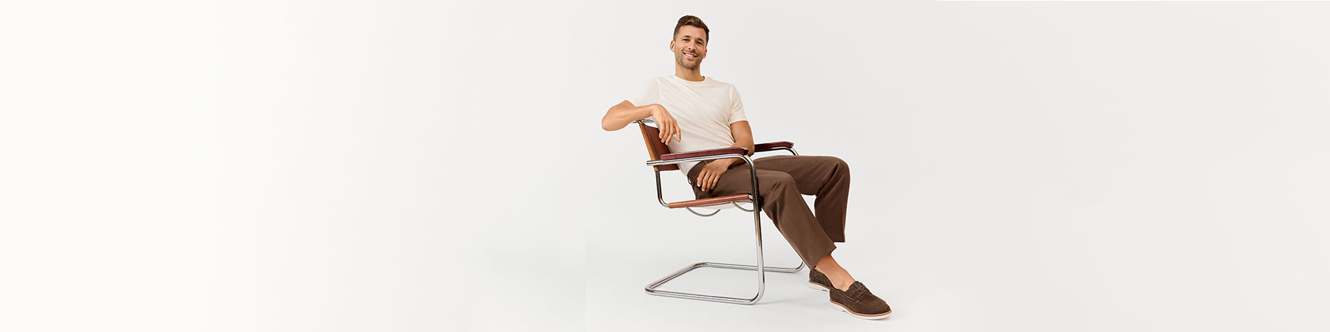 Person in a white T-shirt and brown trousers sits relaxed in a modern chair in front of a light background.