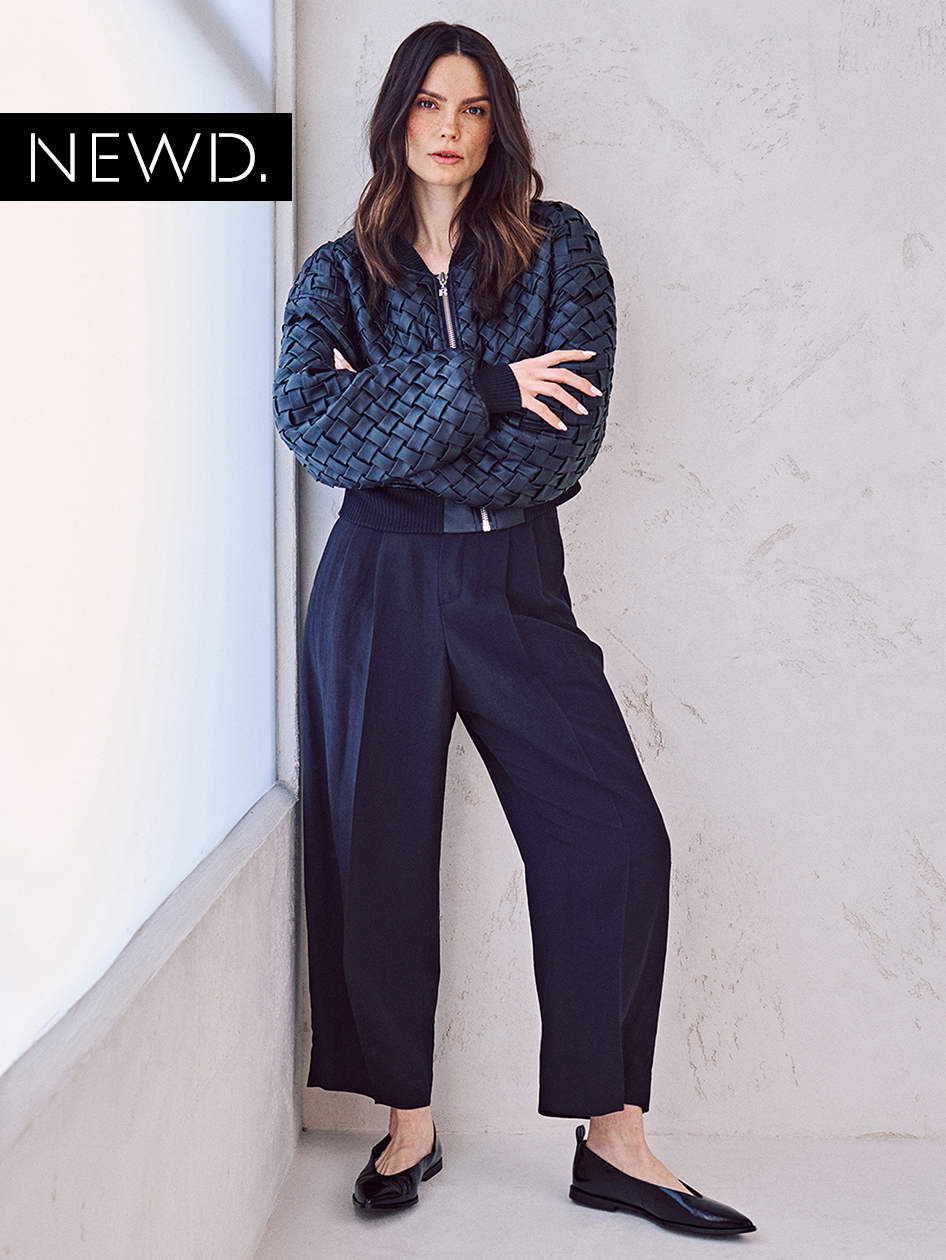 Model in marineblauw bomberjack met geweven patroon en wijde zwarte broek, met platte zwarte schoenen voor een witte muur