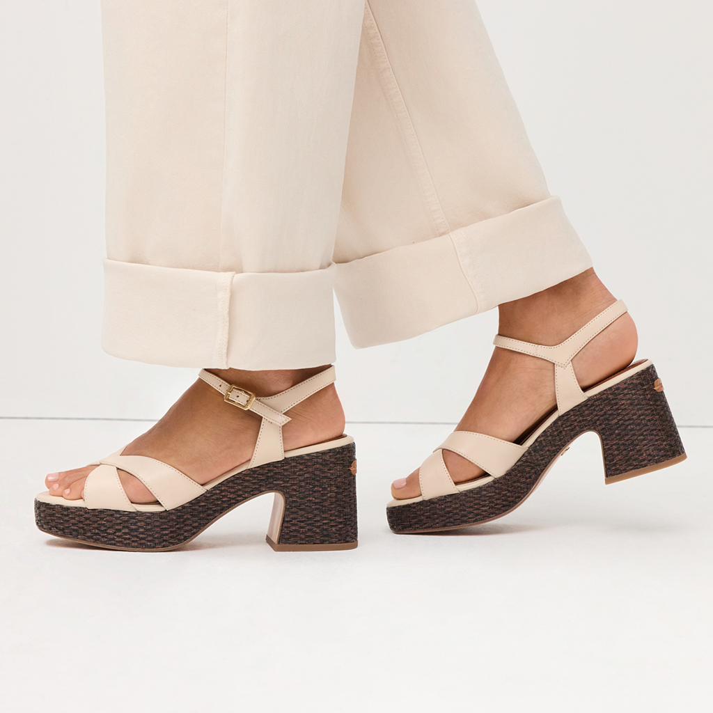 Beige plateau-sandaler med flettet m&oslash;rk h&aelig;l, stylet med hvide bukser mod lys baggrund.