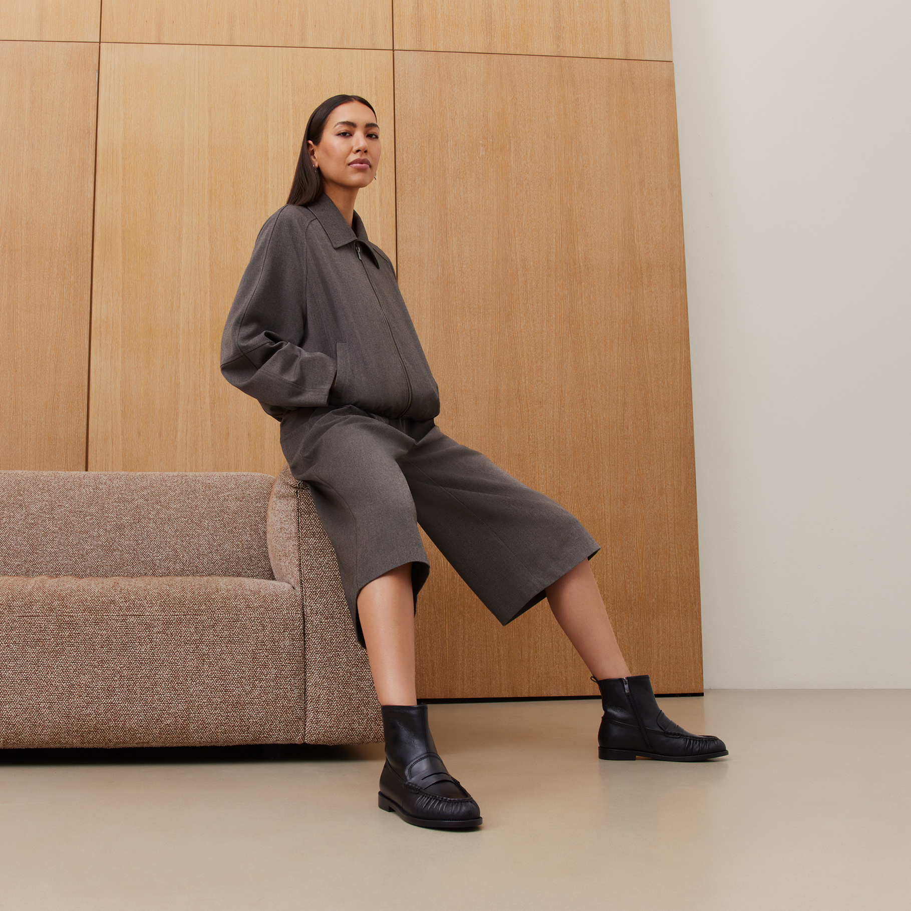 Frau in brauner Jacke und Culotte-Hose sitzt auf beigem Sofa, tr&auml;gt schwarze Ankle-Boots, vor Holzwandpaneelen.