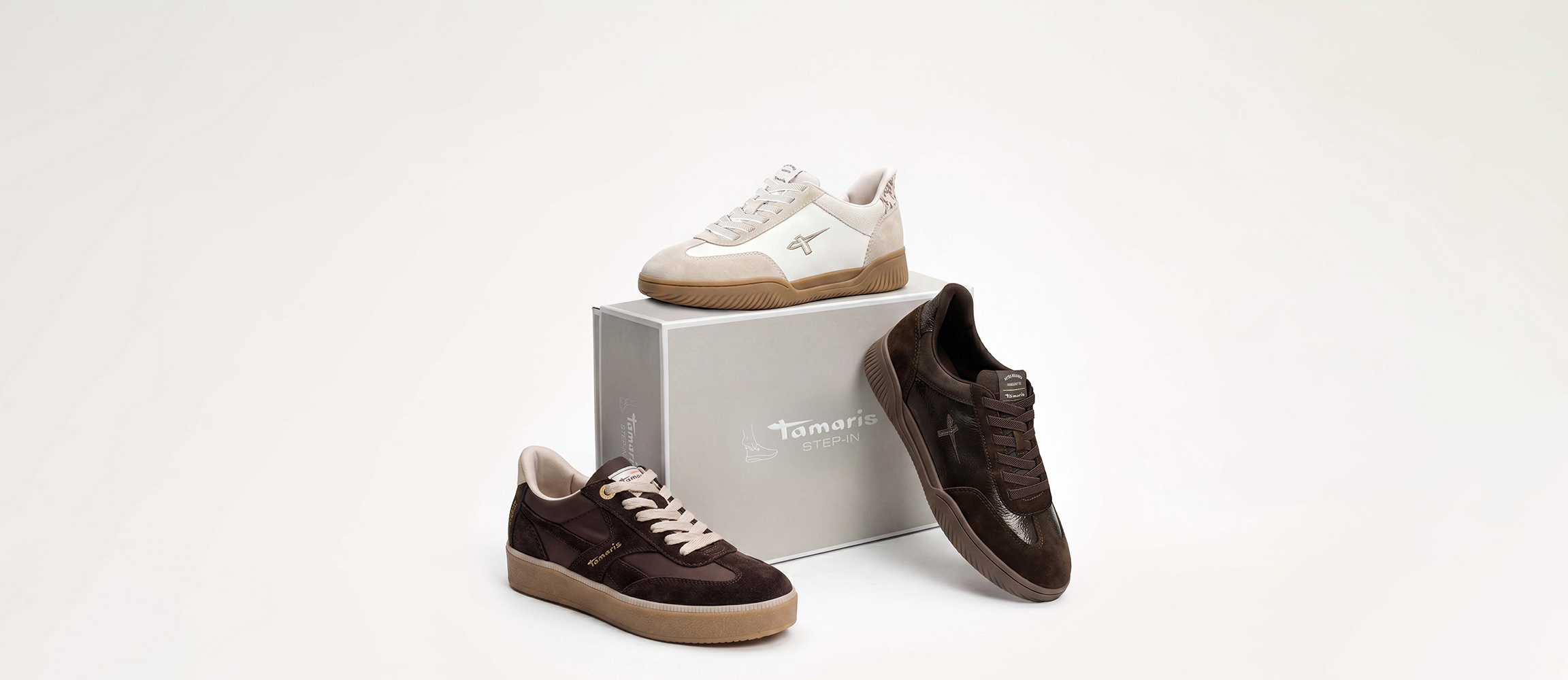 Tamaris-sneakers in wit en bruin, gerangschikt rond een grijze Tamaris Step-In schoenendoos.