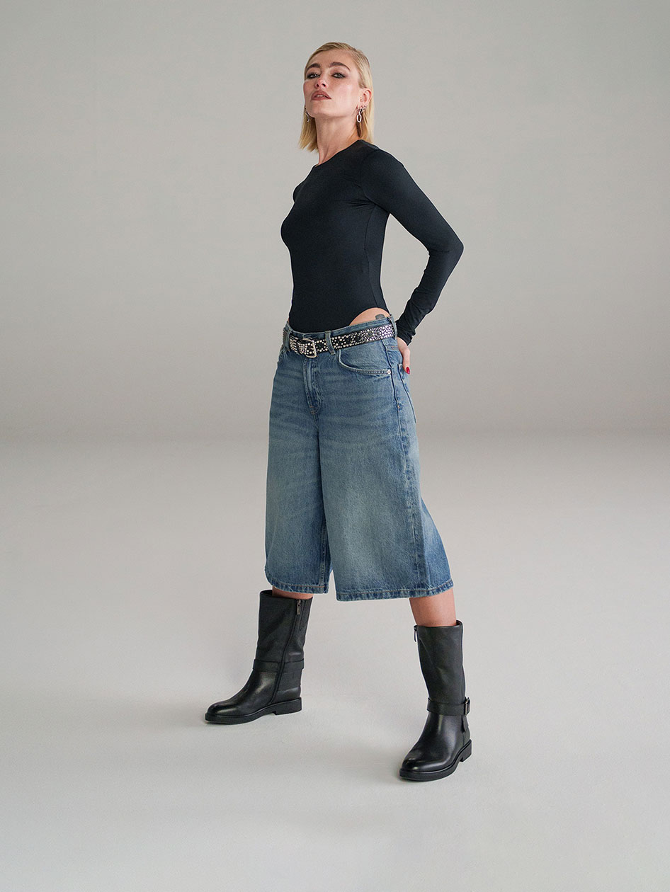 Une femme v&ecirc;tue d'un t-shirt noir &agrave; manches longues, d'un jean culotte large, d'une ceinture &agrave; clous et de bottes montantes noires pose devant un fond gris.
