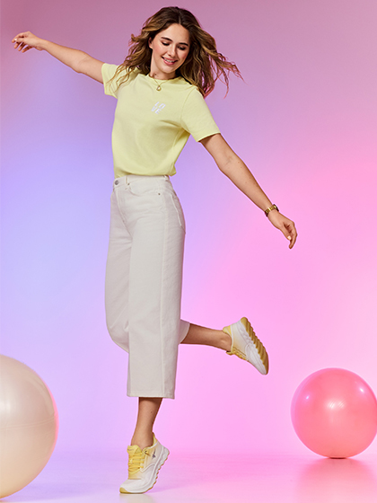Eine Frau in gelbem T-Shirt und beiger Jeans springt fr&ouml;hlich vor rosa-blauem Hintergrund mit schwebenden Luftballons.