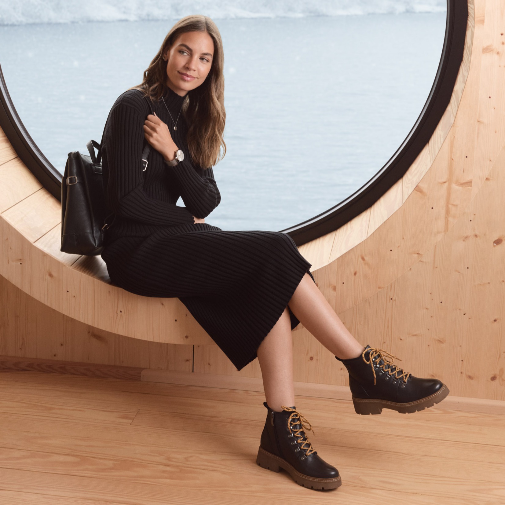 La modella in abito nero lavorato a maglia e robusti stivali stringati siede rilassata su una panca di legno di design dalle forme geometriche.