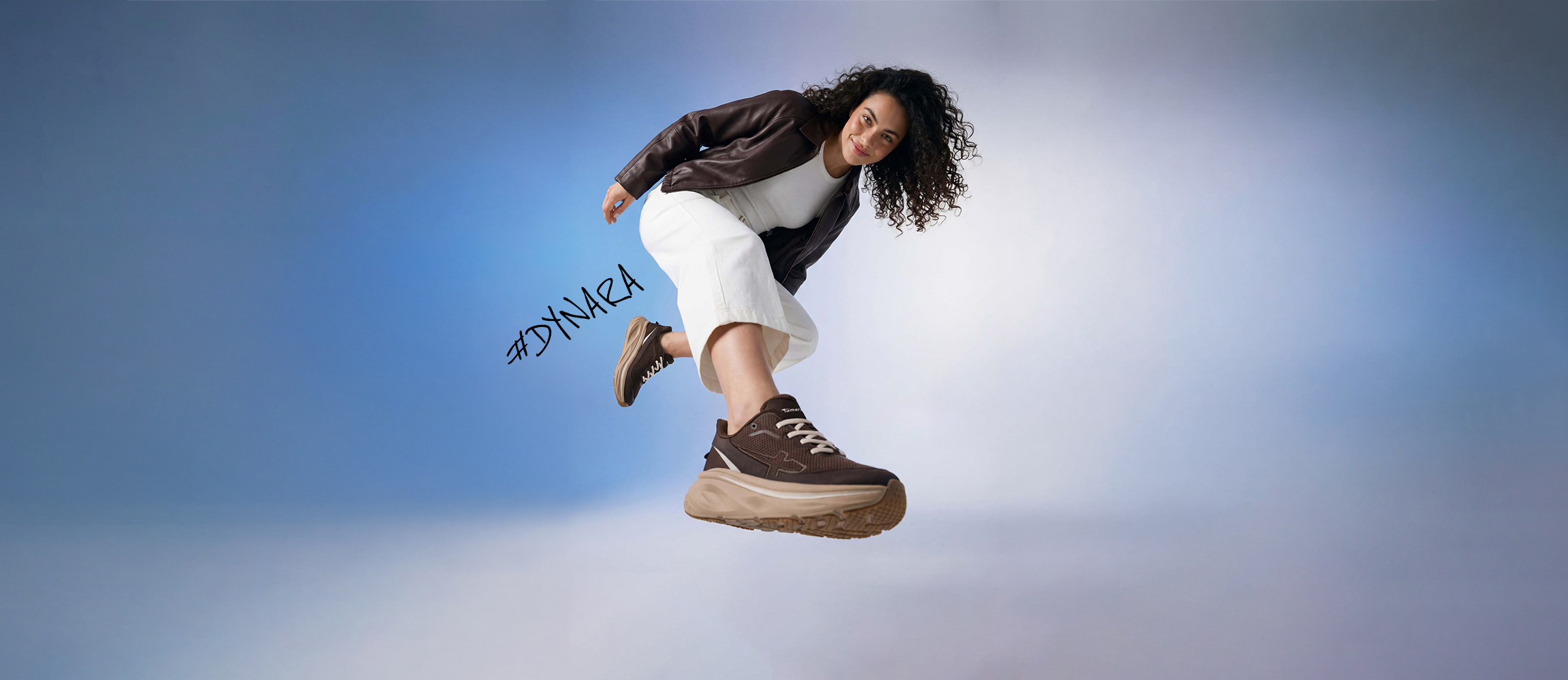 Woman with curly hair jumping in a brown leather jacket, white trousers and brown Tamaris trainers against a blue background. #DYNARA