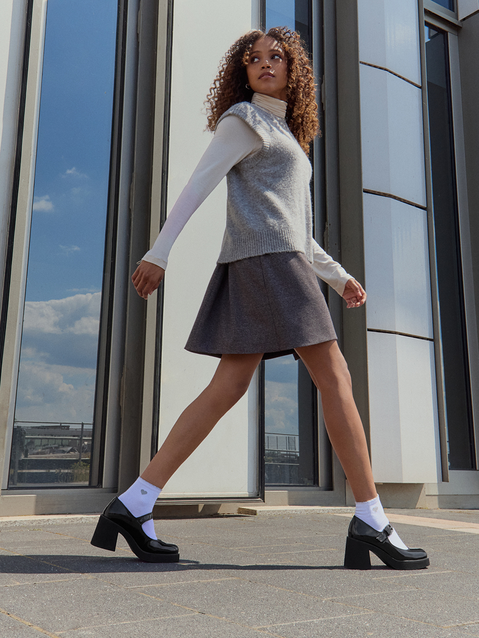 Fashionable autumn outfit with a gray vest, white turtleneck, pleated skirt and black Mary Jane shoes in front of a large window