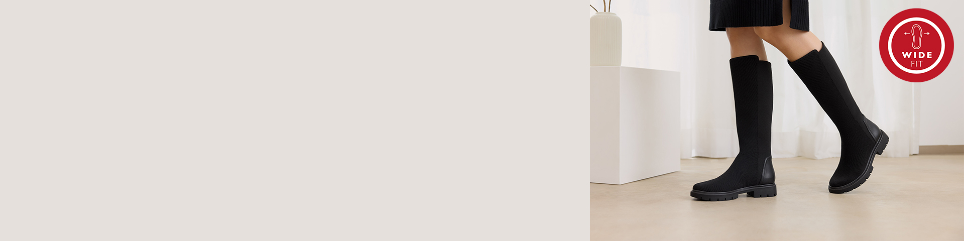 Black knee-high boots with a sturdy sole and wide-fit logo, worn with black knee socks on a light floor.